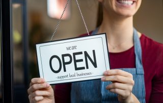 Small business owner smiling while turning the sign for the reopening of the place after the quarantine due to covid-19. Happy businesswoman standing at her restaurant or coffee shop gate with open signboard. Close up of woman"u2019s hands holding sign now we are open support local business.